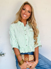 Woman wearing a green and white striped shirt and blue jeans, holding a brown clutch against a white wall.