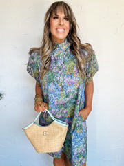 Woman wearing a floral dress holding a straw bag against a white wall