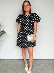 Woman wearing a black polka dot dress with white shoes against a white wall.