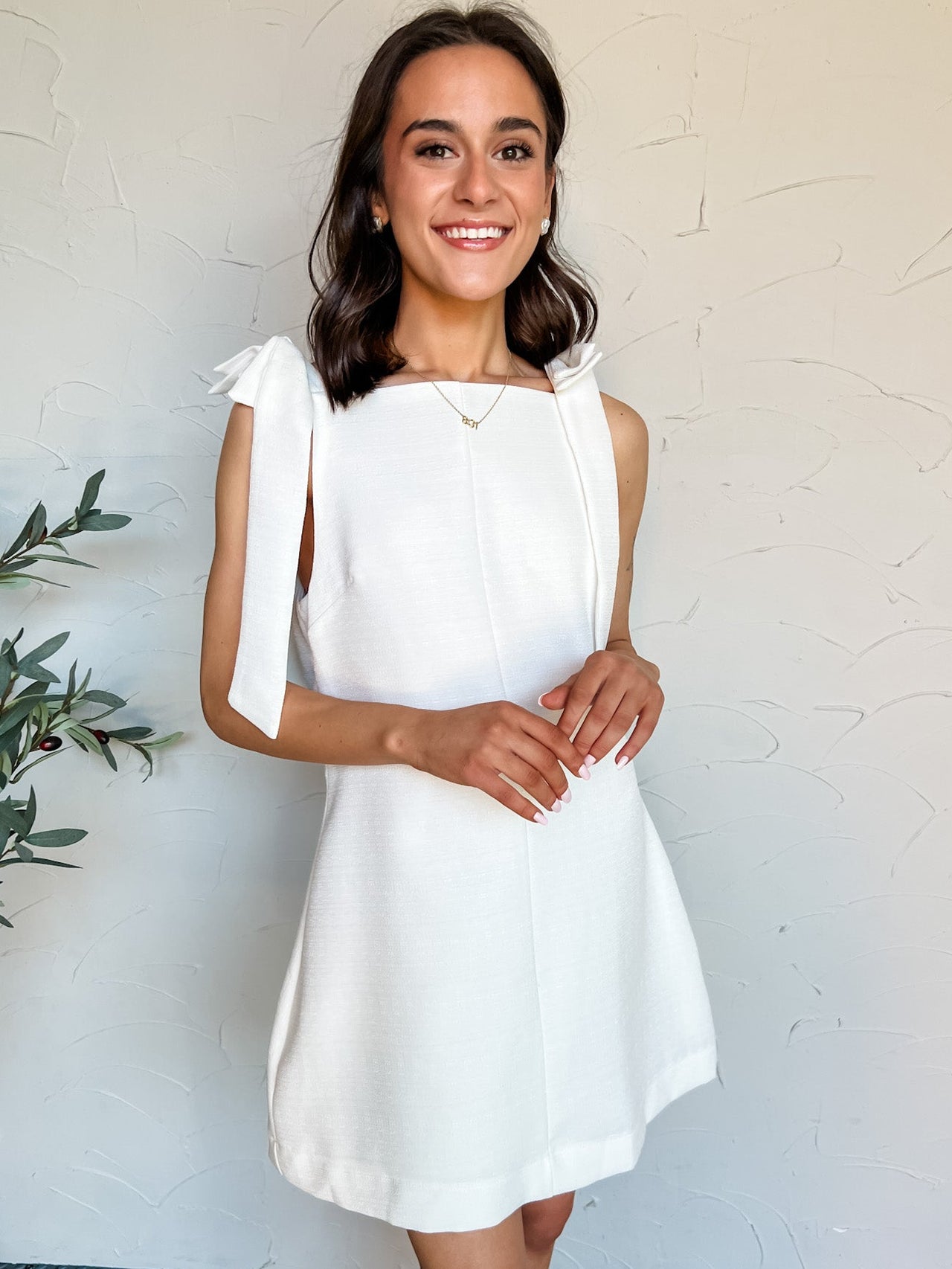 Woman wearing a white dress with bow straps against a plain wall.