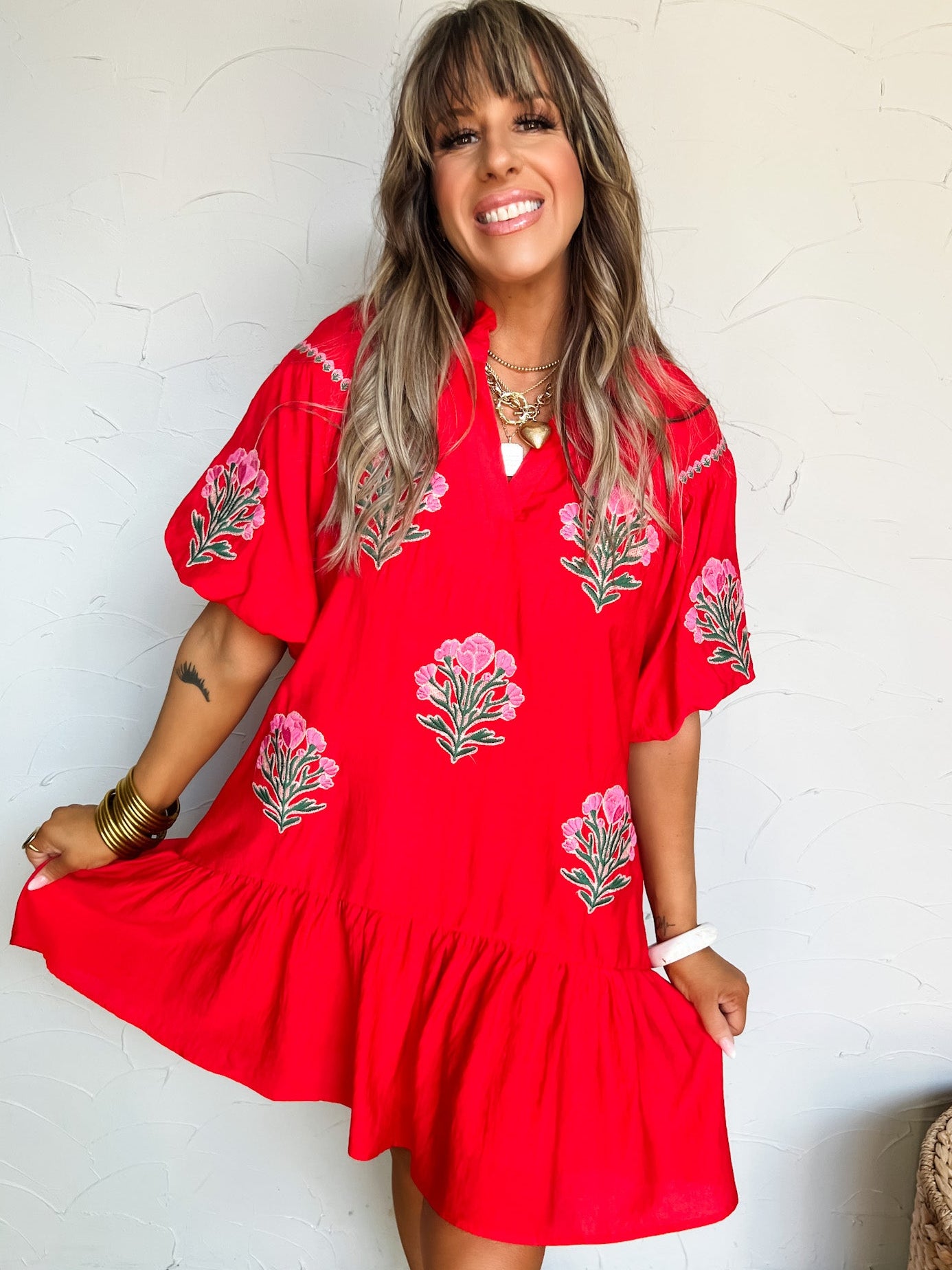 Woman wearing a red dress with floral embroidery against a white wall.
