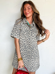 Woman wearing a leopard print dress holding a red handbag against a white wall.