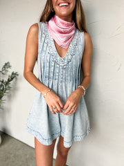 Woman wearing a light blue denim dress with a pink scarf against a white wall.