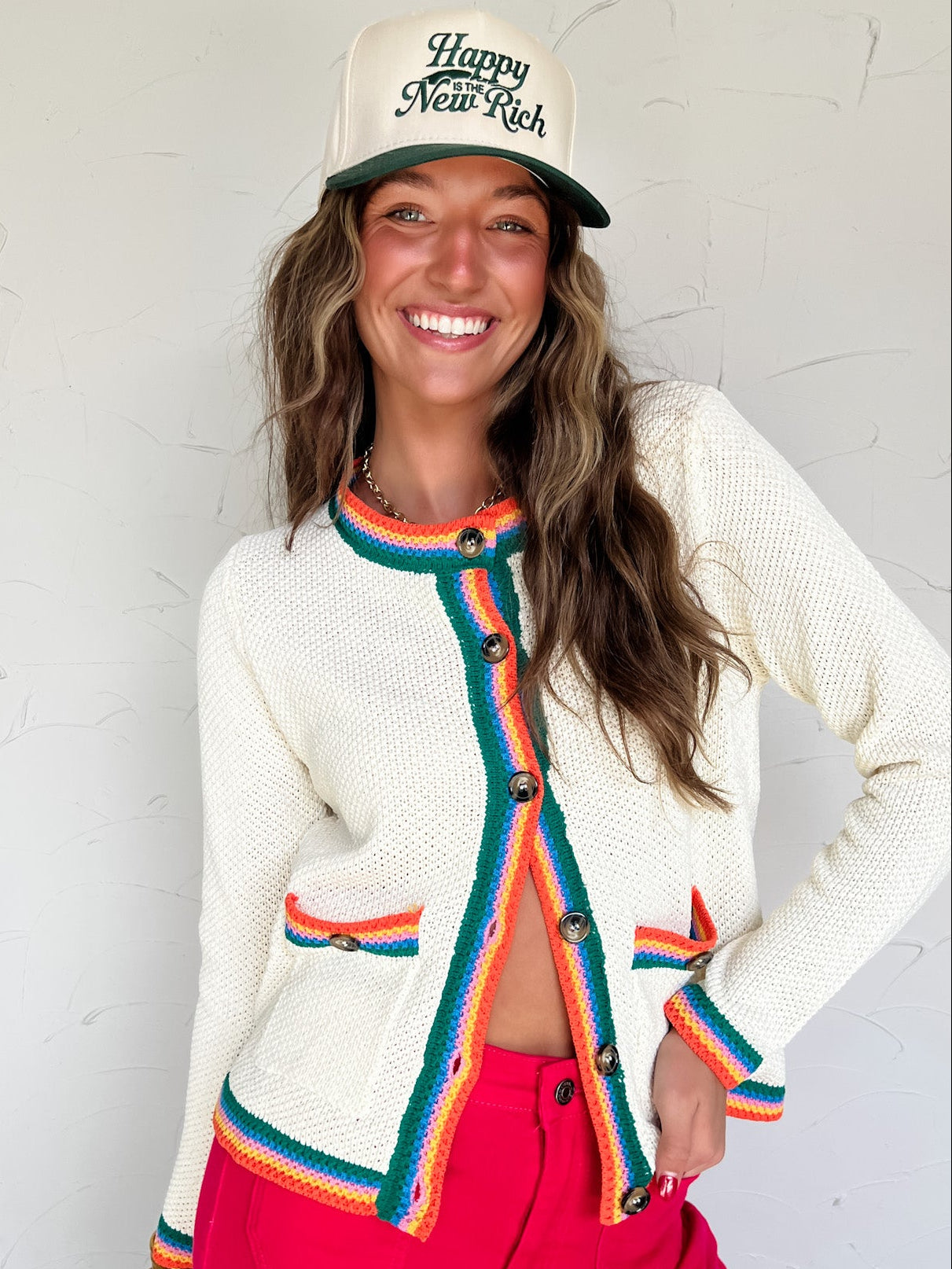 Woman wearing a white cardigan with colorful buttons, pink pants, and a cap with text against a light background