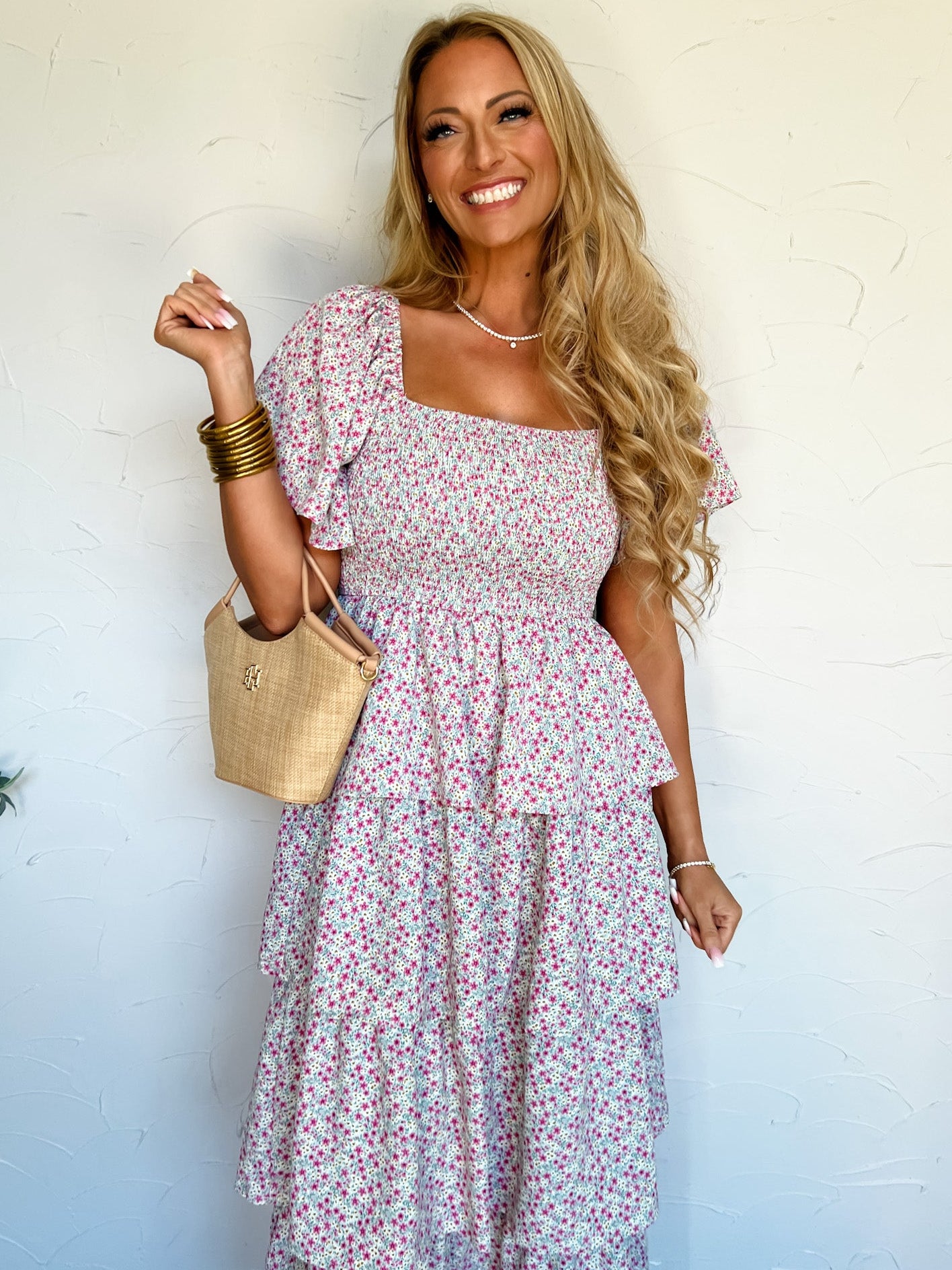 Woman wearing a floral dress holding a straw bag against a white wall.
