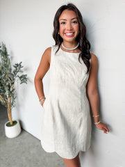 A medium shot of a smiling woman with long dark hair leaning against a textured white wall, wearing a natural cream-colored sleeveless mini dress. The dress is crafted from a woven linen fabric and features a high crew neckline, vertical princess seams along the front for a tailored silhouette, functional side pockets, and a scalloped hemline. She is styled with a classic pearl strand necklace, stacked gold and pearl bracelets on both wrists, and stands next to a small faux olive tree in a white pot.