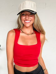 A woman wearing a bright red square neck crop tank top with a black trucker hat and black pants.