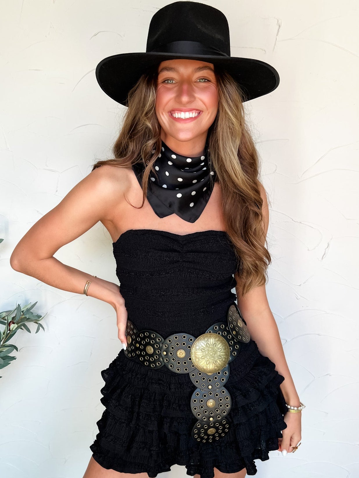 Woman wearing a black dress with ruffles and, a black hat, and white boots indoors.