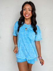 A mid-shot photograph of a smiling brunette woman posing against a textured white wall. She is wearing an oversized crewneck t-shirt made from a 100% cotton fabric with a light blue mineral-wash finish, featuring a left chest patch pocket, dropped shoulders, short cuffed sleeves, and a raw-edge split hemline.