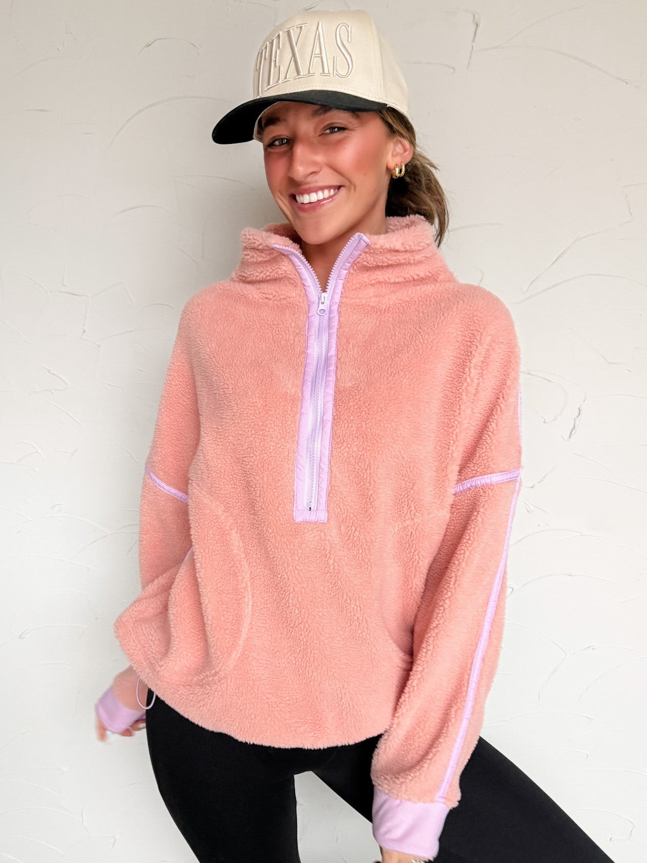 Person wearing a pink fleece pullover with purple detail with a zipper, black leggings, and a beige cap against a white background.