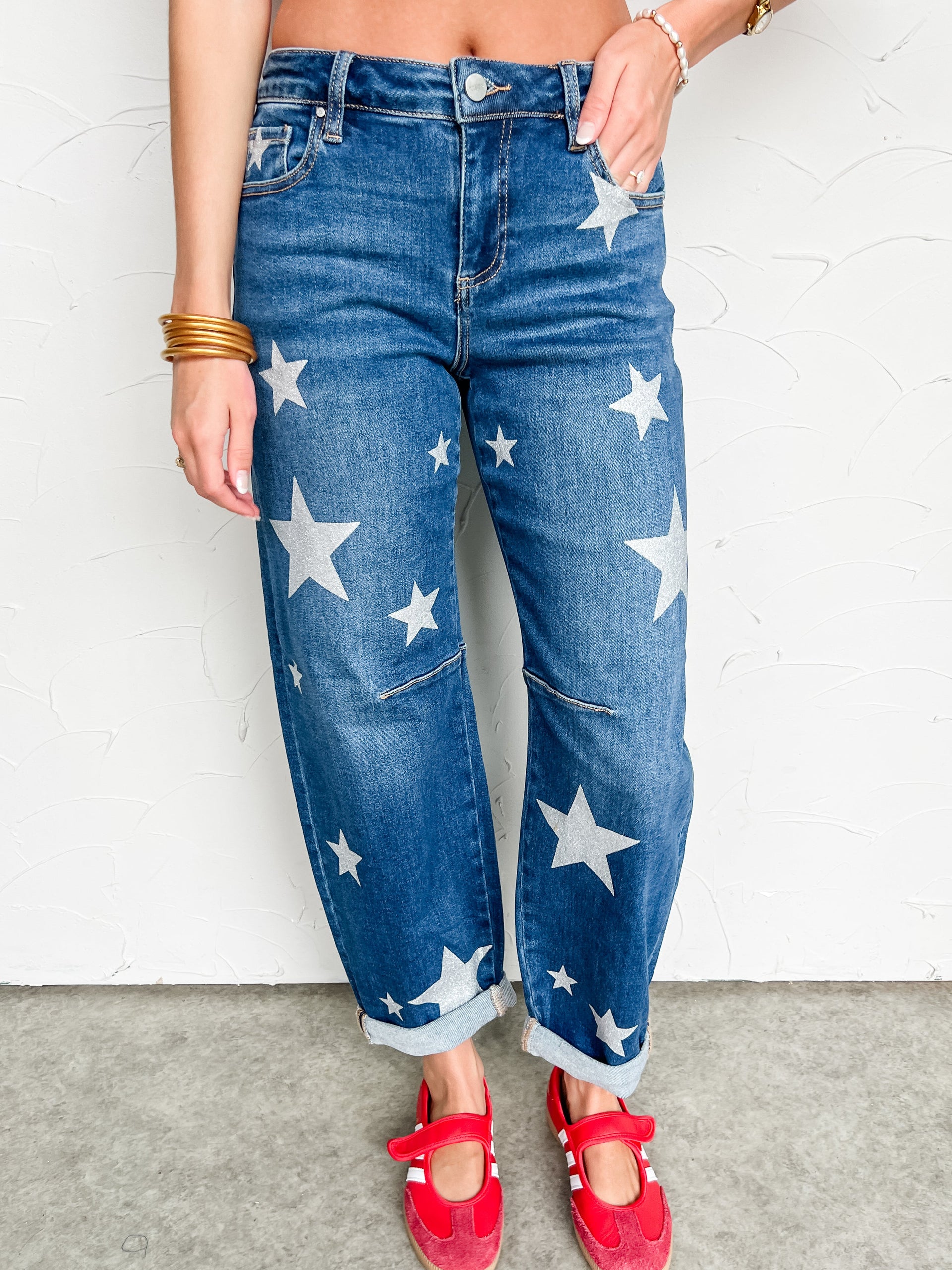 Person wearing blue jeans with silver star patterns and red shoes against a white wall.