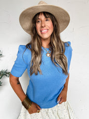 Woman wearing a blue sweater and straw hat against a white wall
