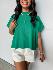 A close-up photograph from the chin down of a smiling brunette woman posing against a textured white wall. She is wearing an oversized mock neck top made from a ribbed 72% viscose and 28% polyester blend fabric in a vibrant green colorway, featuring a vertical ribbed pattern and relaxed short sleeves.