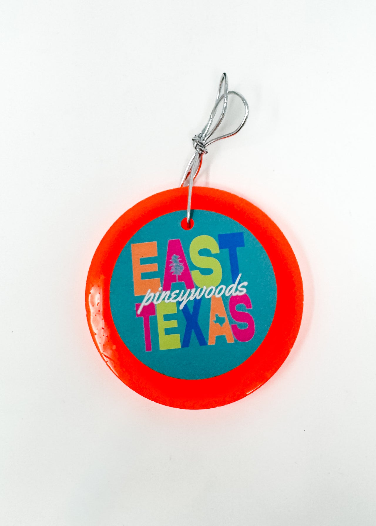 This "East Texas Pineywoods" car freshie features a colorful, round design with a bold retro-style graphic, a small pine tree detail, and a mini Texas state silhouette, all framed by a smooth, circular outer border. It comes ready to hang from your rearview mirror with an attached silver string. Colors and scents vary.