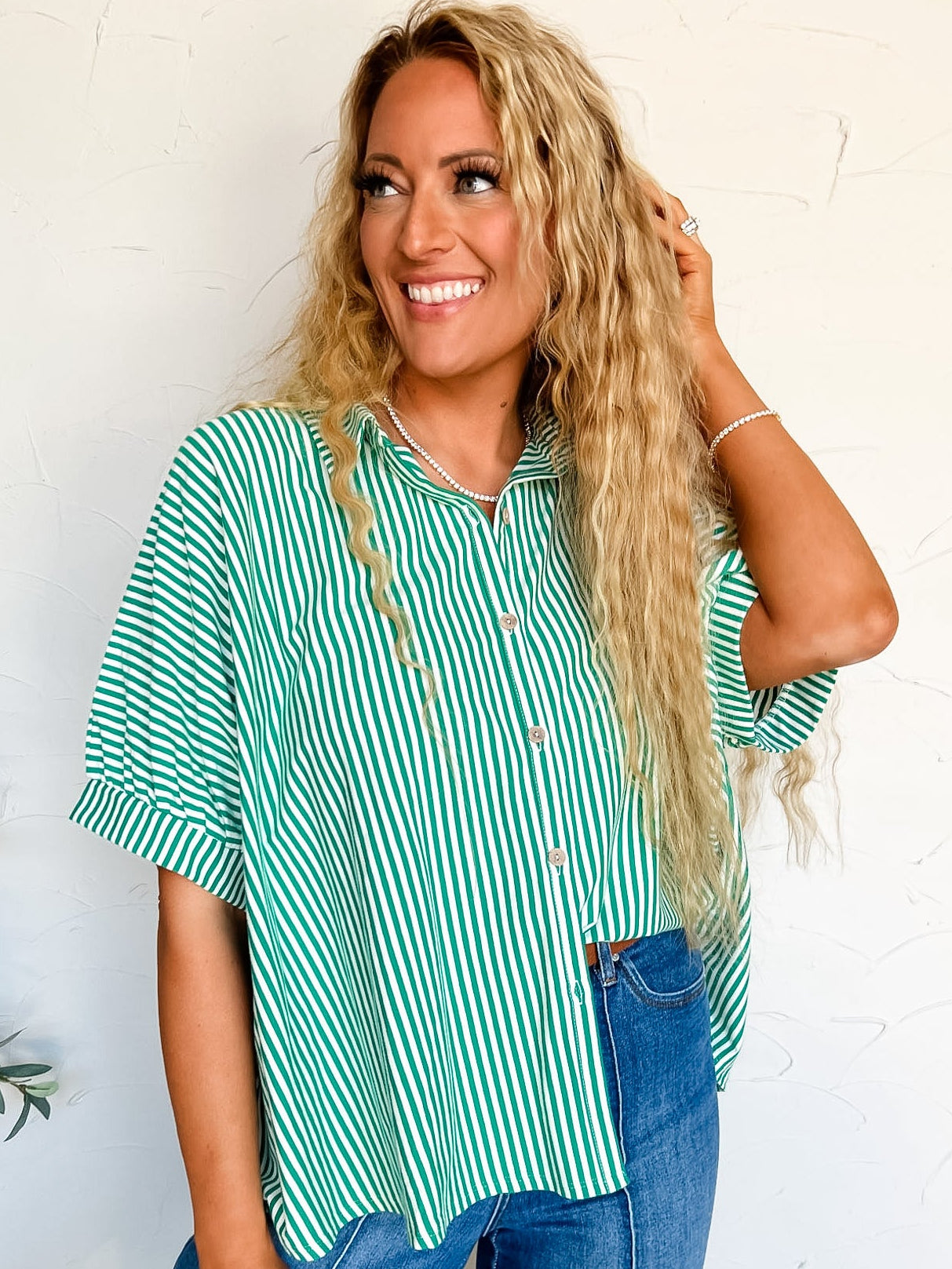Woman wearing a green and white striped button up shirt and blue jeans against a white wall.