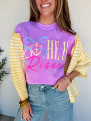 Person wearing a purple shirt with 'HE RISES' text, yellow striped cardigan, and blue jeans against a white wall.