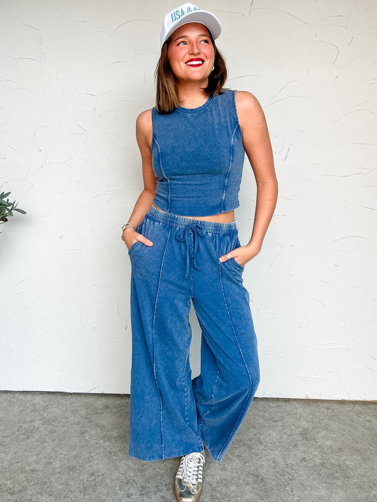 Woman wearing a blue acid washed set outfit with a white cap against a white wall.