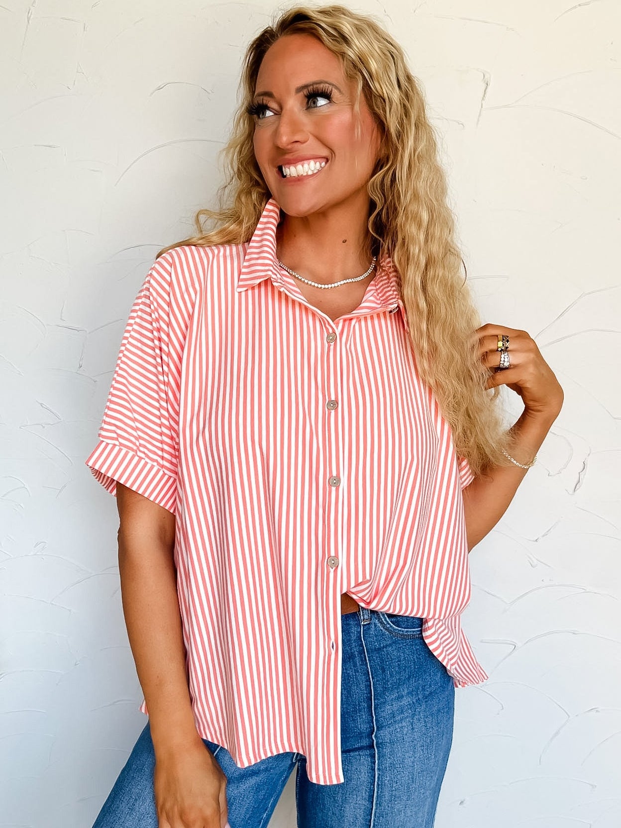 Woman wearing a coral and white striped button up shirt and blue jeans against a white wall.