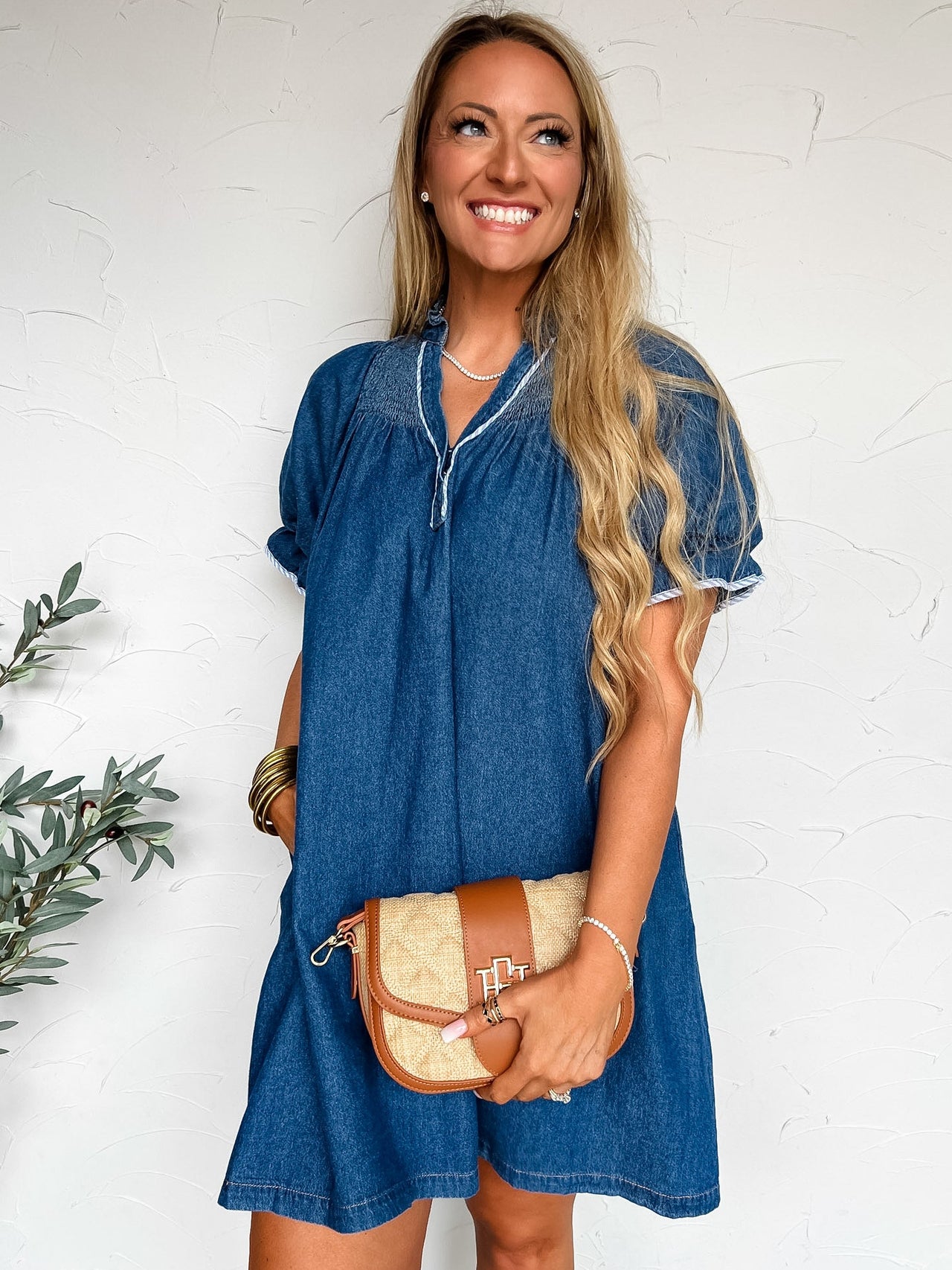 Woman wearing a blue denim dress holding a beige handbag against a white background
