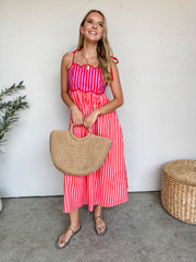 A woman smiling against a textured off-white wall, wearing a lined 100% cotton midi dress featuring a vibrant striped pattern in shades of pink and red. The dress showcases a structured sweetheart neckline with a distinctive scalloped bustline paneled in vertical purple and pink stripes, accented by solid red tie-shoulder straps. The flowing A-line skirt drops into a midi length with vertical red and pink stripes. She is styled with a gold link chain heart necklace and metallic strappy flat sandals, standin