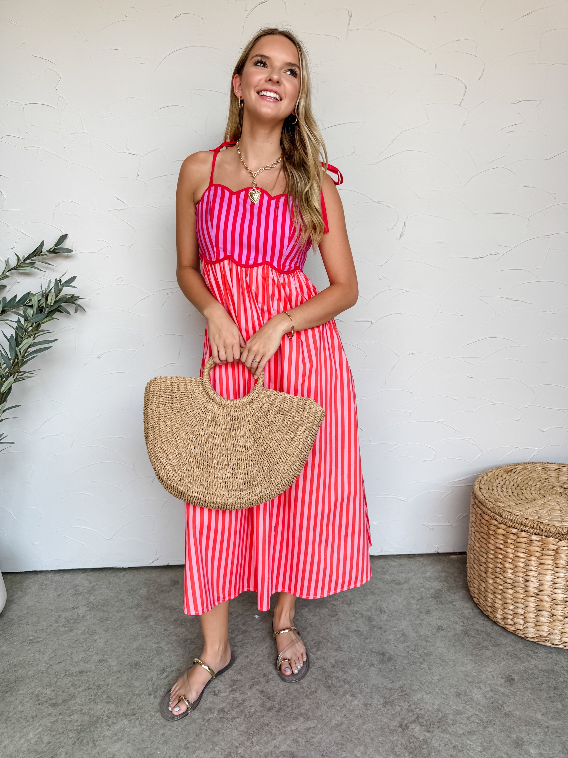 A woman smiling against a textured off-white wall, wearing a lined 100% cotton midi dress featuring a vibrant striped pattern in shades of pink and red. The dress showcases a structured sweetheart neckline with a distinctive scalloped bustline paneled in vertical purple and pink stripes, accented by solid red tie-shoulder straps. The flowing A-line skirt drops into a midi length with vertical red and pink stripes. She is styled with a gold link chain heart necklace and metallic strappy flat sandals, standin
