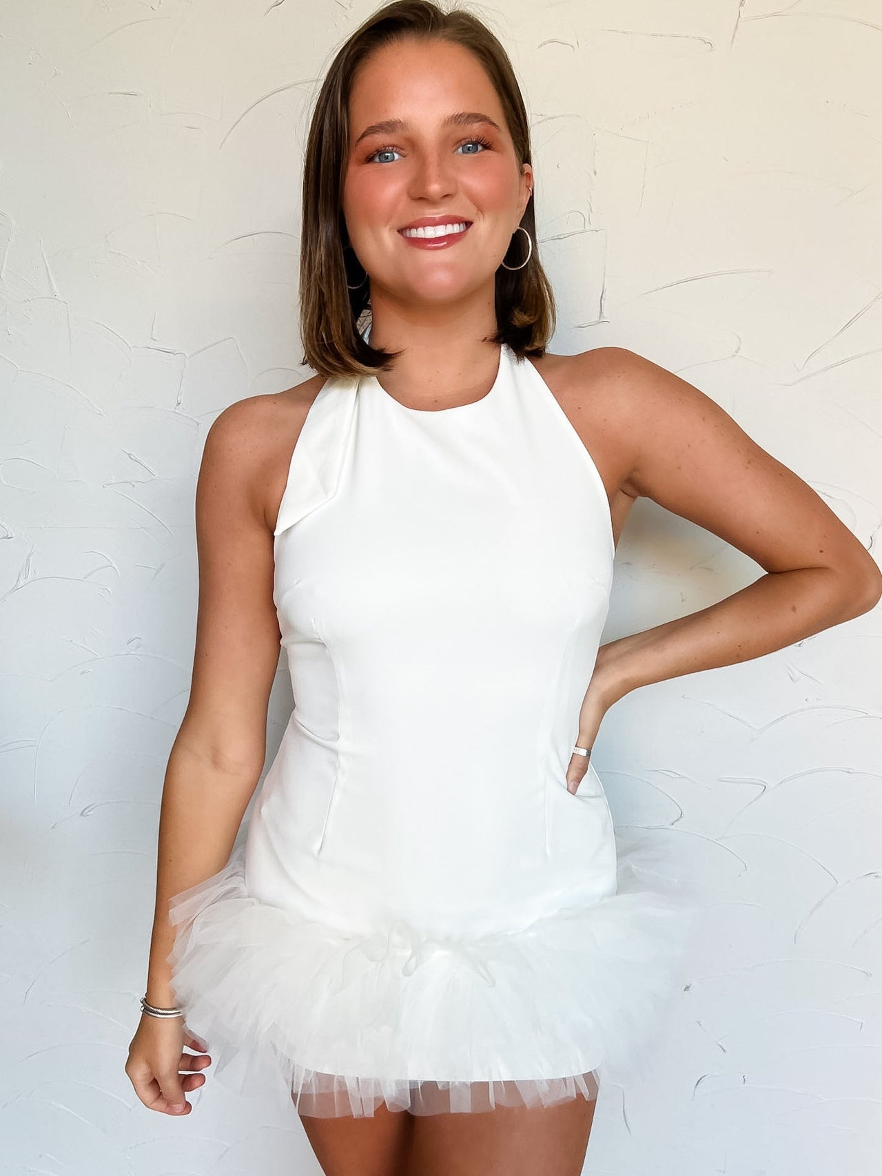 Woman wearing a fitted white dress with ruffle tulle on hem against a light background