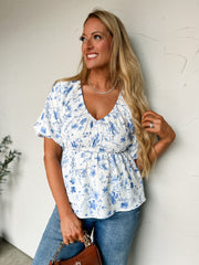 Woman wearing a blue floral top and jeans against a white wall.