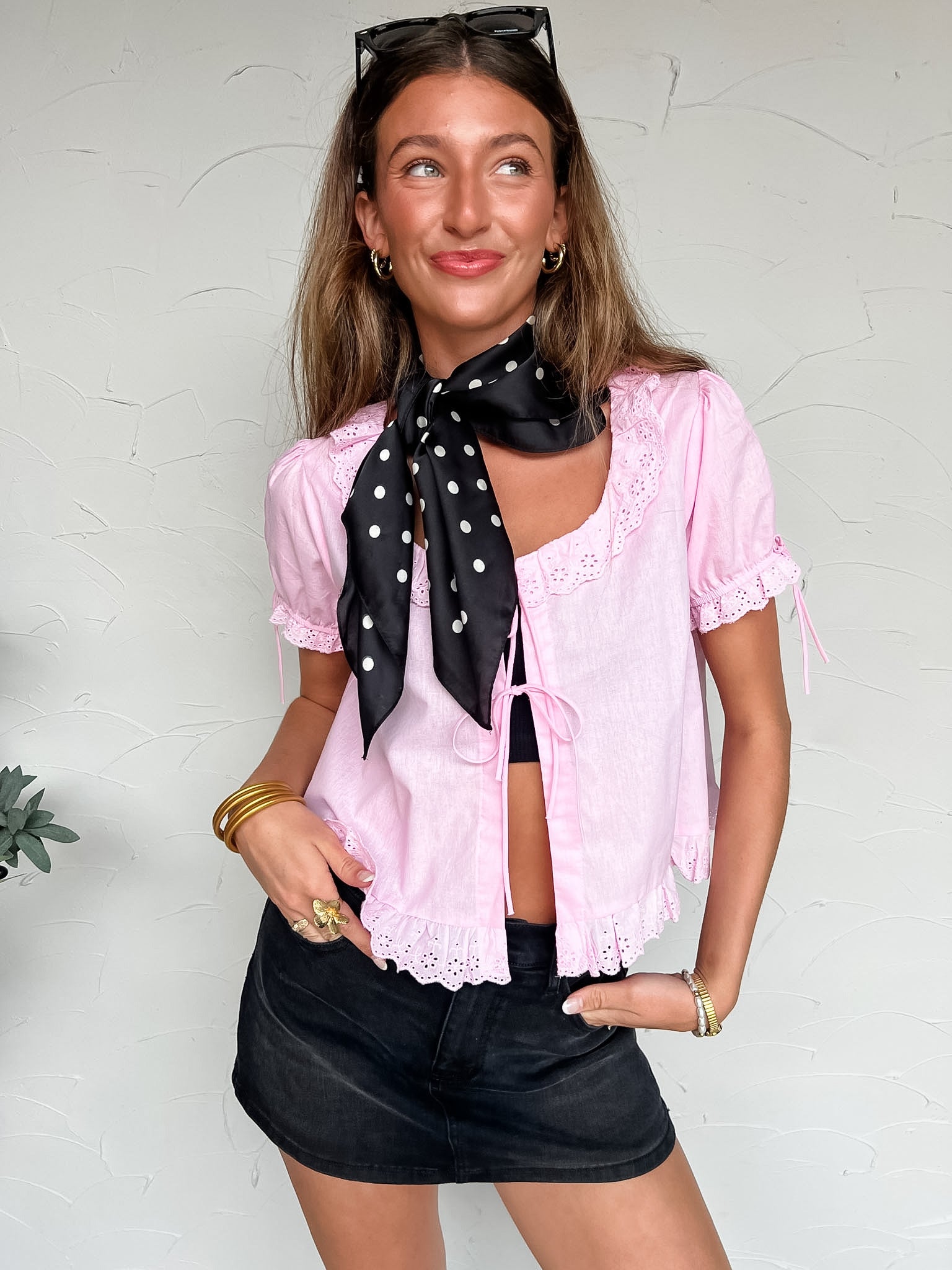 Woman wearing a pink blouse with lace details and black shorts, standing against a white wall.