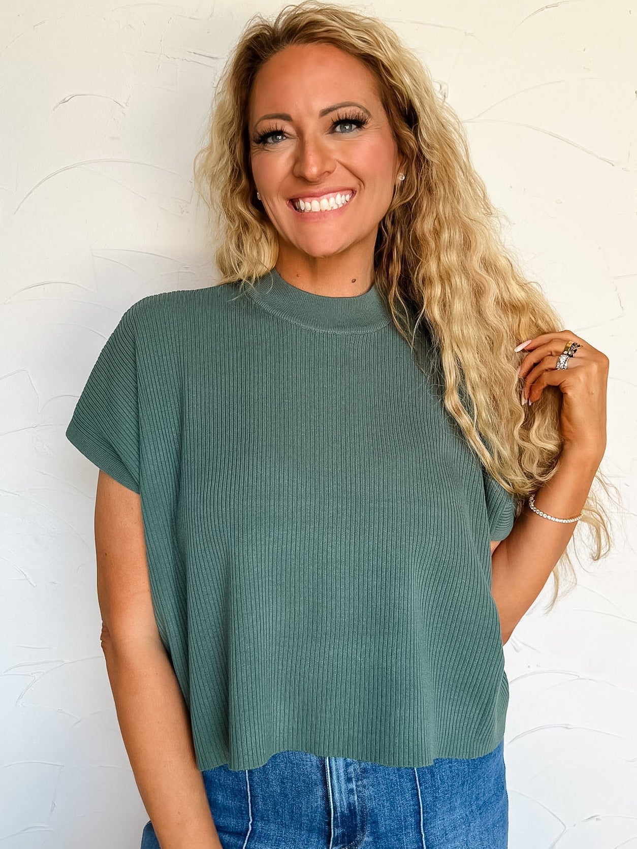 Woman wearing a green ribbed sweater top and blue jeans against a white wall