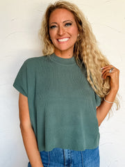 Woman wearing a green ribbed sweater top and blue jeans against a white wall