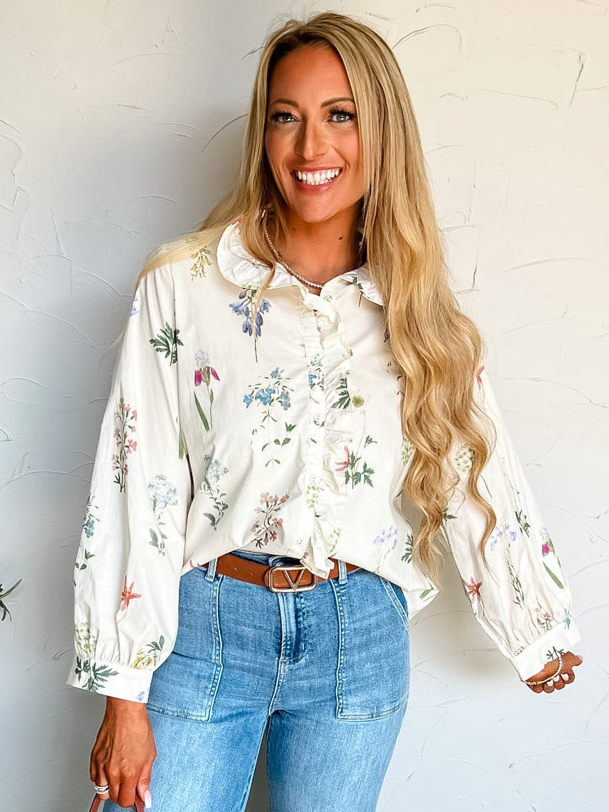 Woman wearing a floral blouse and jeans, holding a straw bag against a white wall.