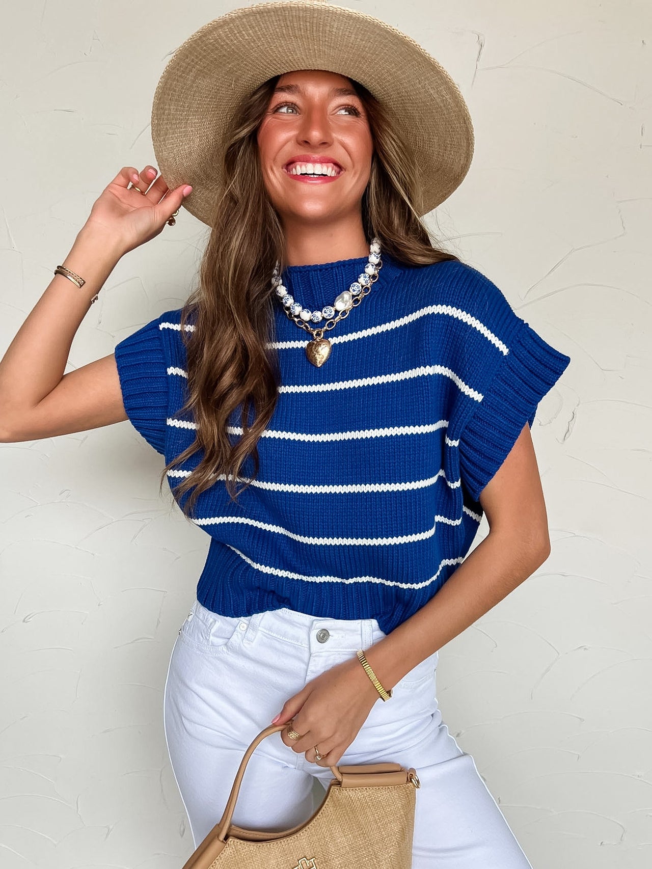 Woman wearing a blue and white striped sweater, white pants, and a straw hat against a light background