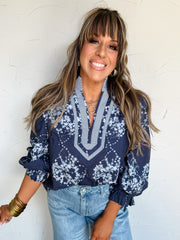Woman wearing a navy blue patterned blouse and light blue jeans against a white wall.
