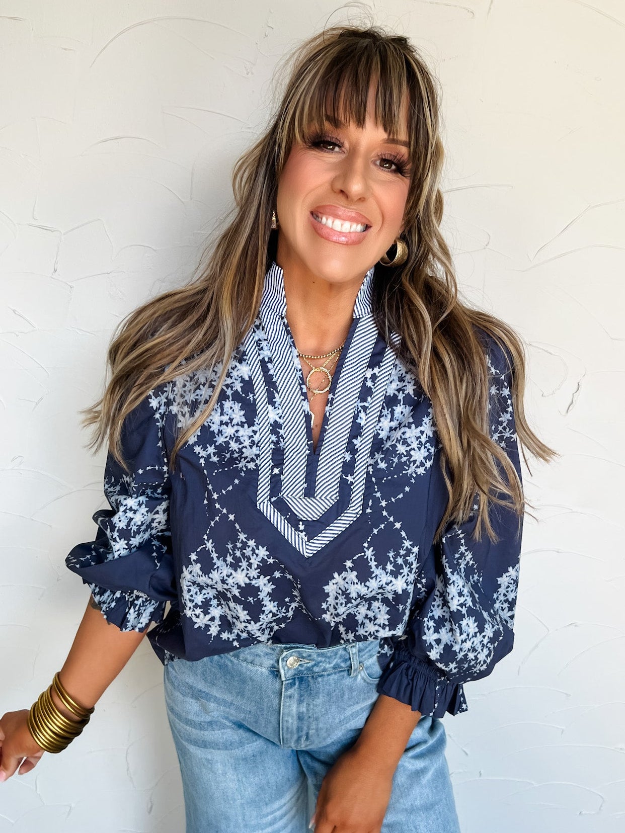 Woman wearing a navy blue patterned blouse and light blue jeans against a white wall.