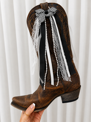 Brown cowboy boot with decorative ribbons and lace against a white curtain background