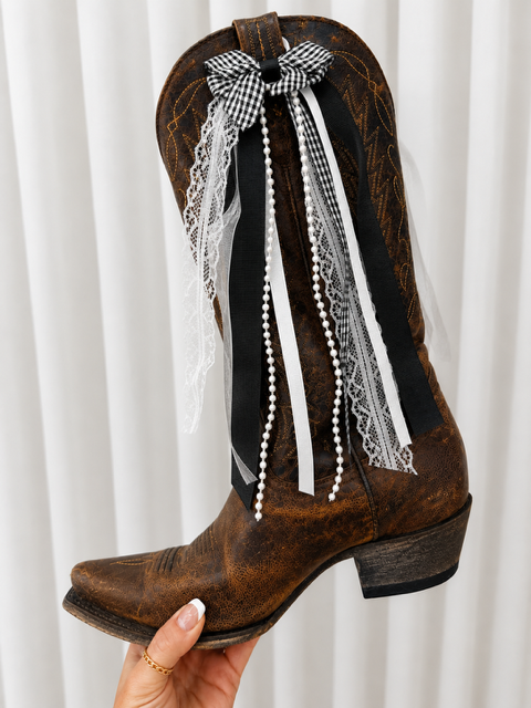 Brown cowboy boot with decorative ribbons and lace against a white curtain background