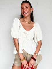 Woman wearing a white blouse and beige shorts against a white wall.