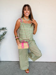A smiling woman poses against an off-white wall wearing a green and pink block-print matching set crafted from 100% cotton. The two-piece set includes a square-neck sleeveless top with button-front detailing and wide straps, paired with coordinating high-waisted, wide-leg pants. She is styled with a chunky mint green necklace, stacked gold bangles with a pink accent bracelet, a straw handbag with bright pink leather trim, and tan flat sandals.
