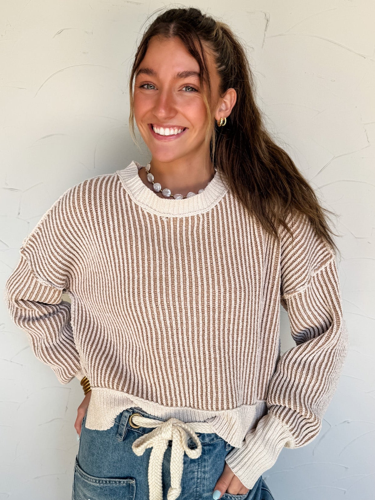 Woman wearing a brown mix striped/ribbed sweater and jeans against a white wall