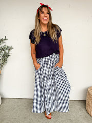 A woman smiling with her hands in her pockets against a textured off-white wall, wearing a one-piece mixed-media maxi dress featuring a bodice crafted from a 50% viscose, 28% polyester, and 22% nylon knit blend. The top portion of the dress is solid navy blue with a round neckline and short sleeves, flowing into an attached, voluminous navy blue and white gingham patterned woven maxi skirt. The skirt is detailed with large, functional front patch pockets. She is styled with a bright red bandana tied as a he