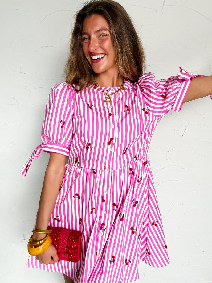 Woman wearing a pink and white striped dress with red cherries and a red clutch against a white background