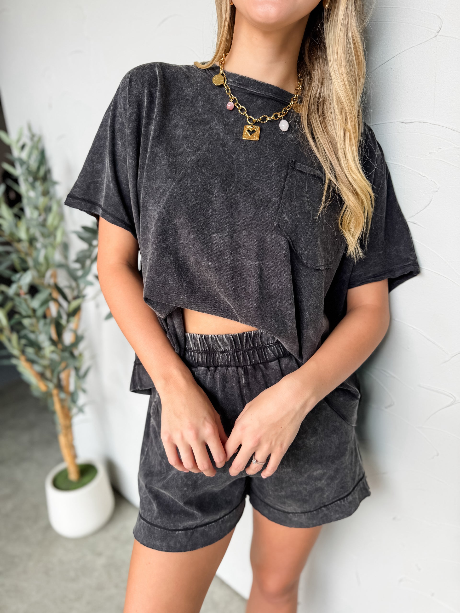 A full-length photo of a smiling blonde woman posing against a white textured wall. She is wearing a charcoal grey acid-wash matching lounge set consisting of an oversized, boxy short-sleeved top with a single chest pocket and matching high-waisted shorts with an elastic waistband. She is balancing playfully on one leg with her left hand touching her hair, wearing chunky white sneakers and a layered gold chain necklace. A small potted olive tree is visible on the left.
