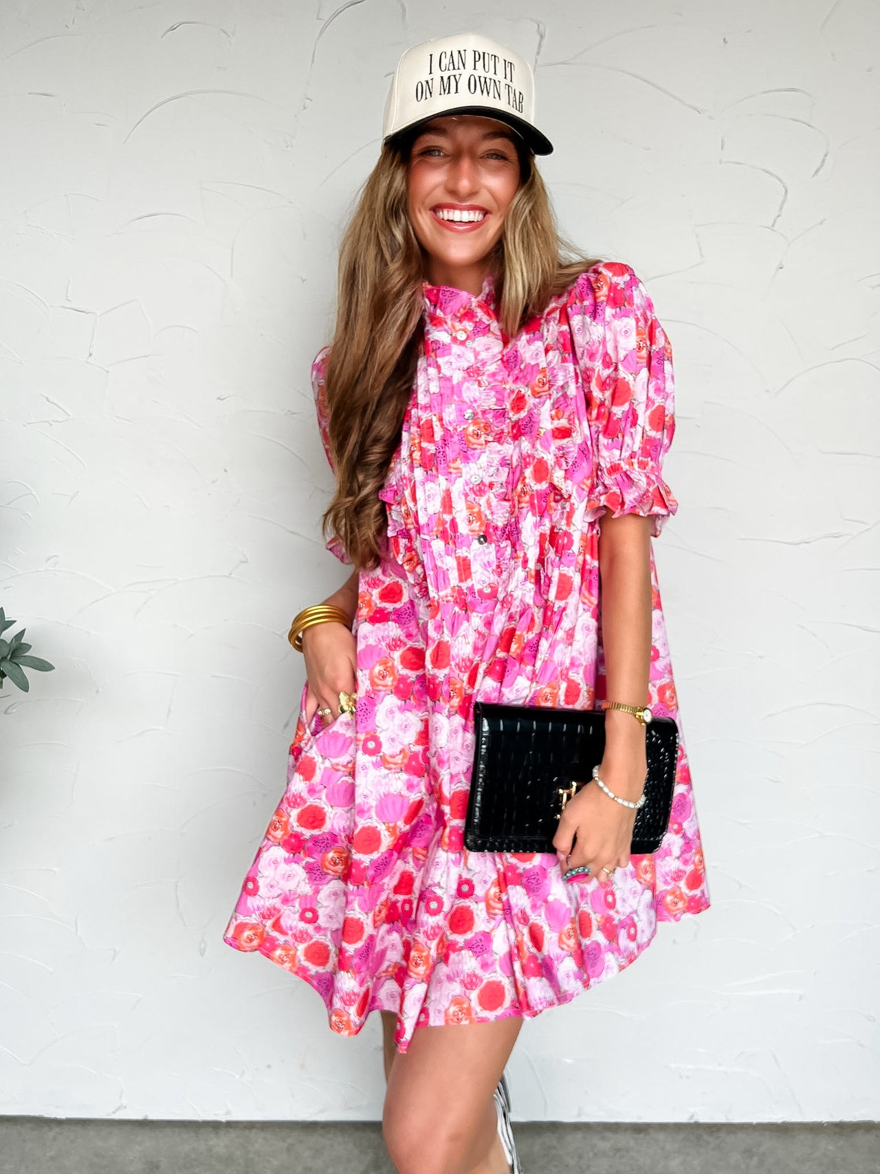 Woman wearing a pink floral dress and white cap with text, holding a black clutch against a white background.