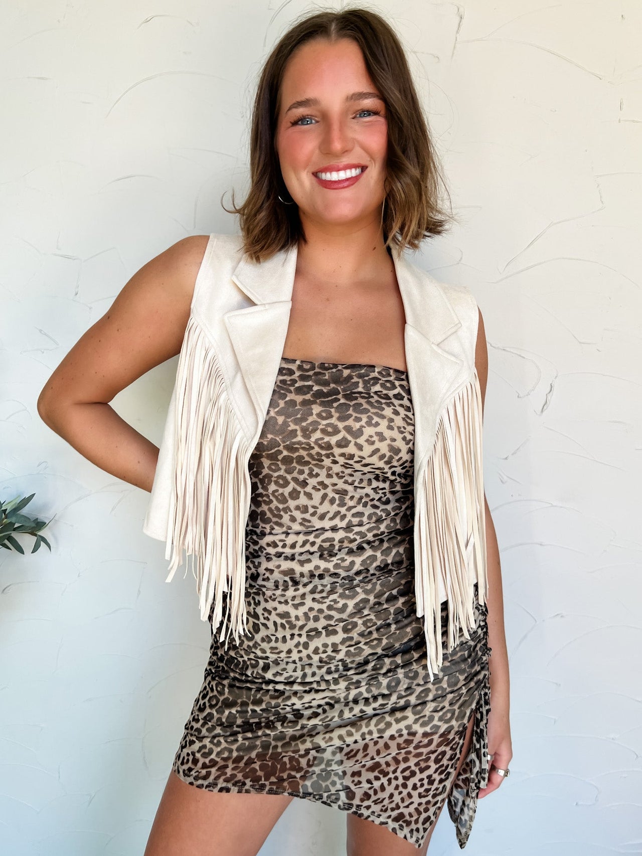 Woman wearing a leopard print dress with a white fringe vest against a white wall.