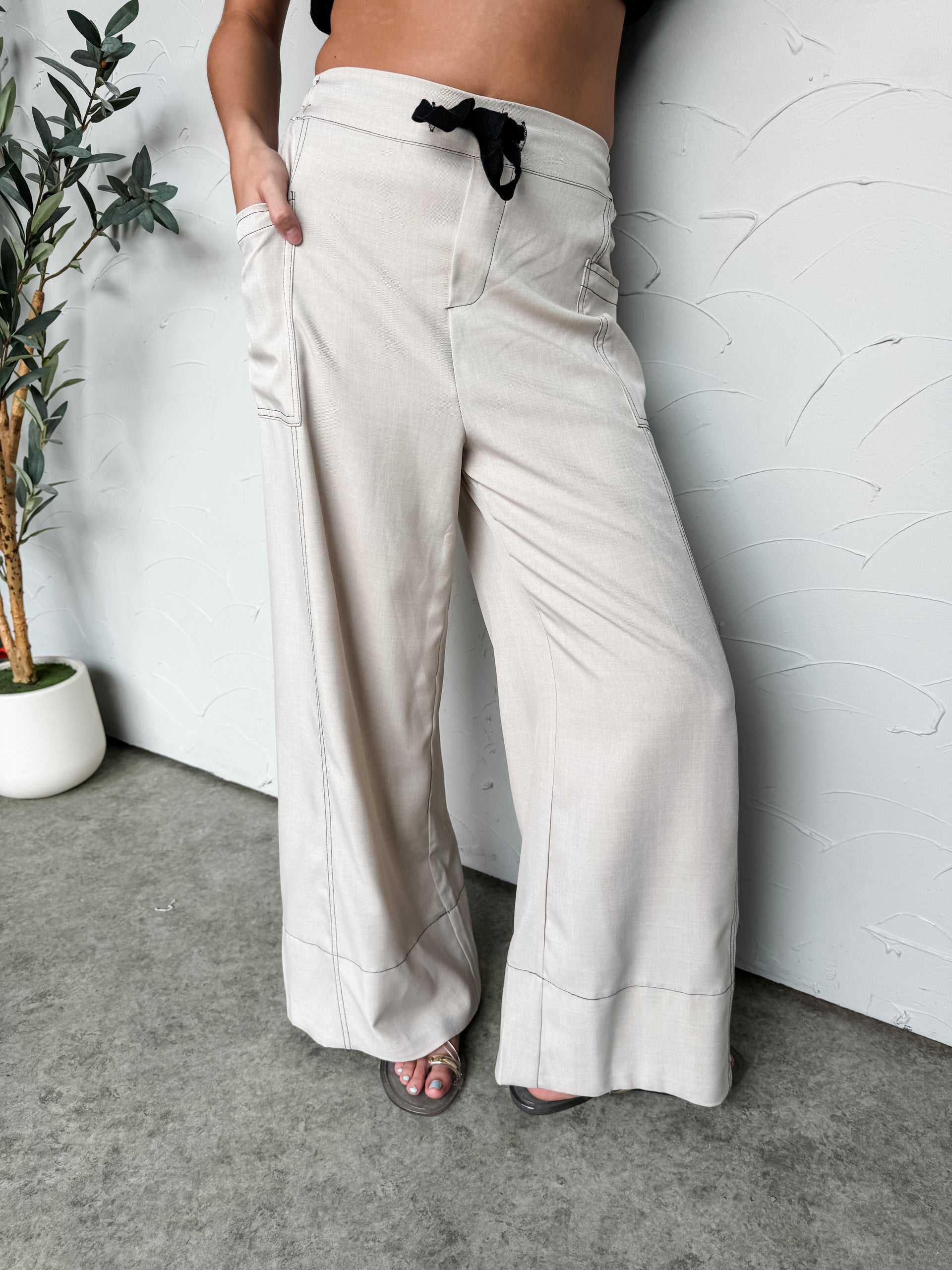 A front-view, cropped photo focusing from the waist down of a person wearing wide-leg trousers in a neutral oatmeal color against a textured white wall. The pants feature a flat front waistband secured by a black velvet drawstring tie, dark contrast topstitching details along the seams, and functional side utility utility-style patch pockets. The person is also wearing a black top that is partially visible, exposing their midriff, and flat sandals that reveal a light blue pedicure.