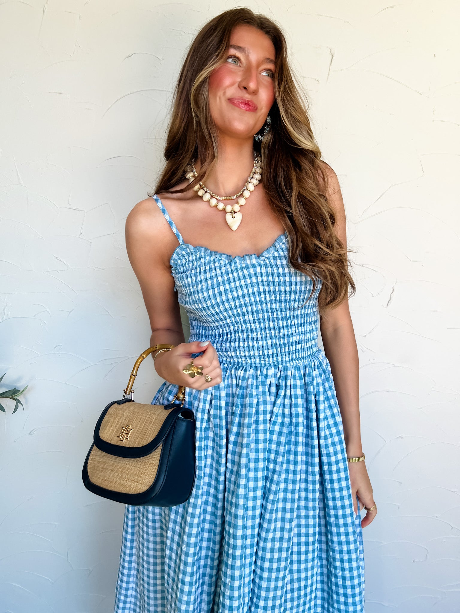 Woman wearing a blue gingham dress holding a handbag against a white wall.