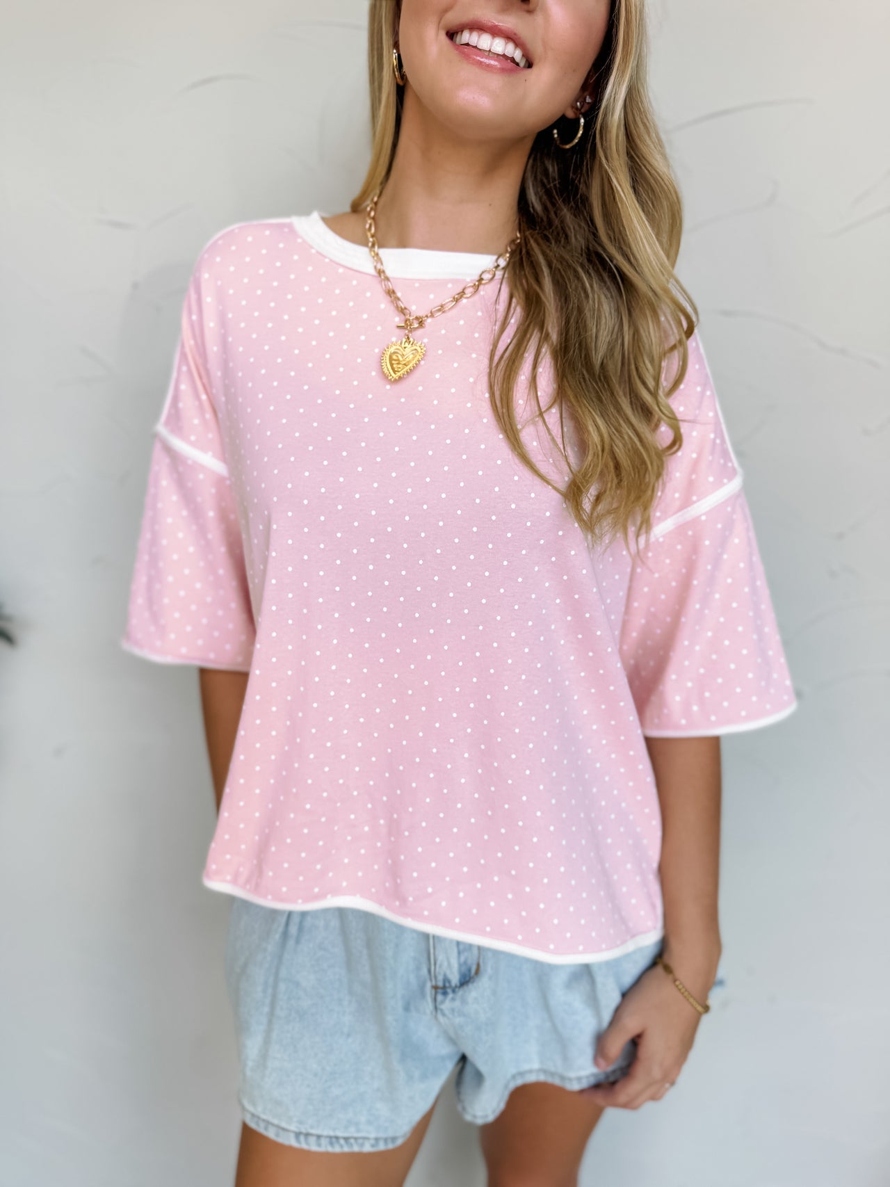 Smiling model wearing a relaxed-fit blush pink top made from a soft cotton-spandex knit fabric. The top features an allover white polka dot print, crew neckline, dropped shoulders, short sleeves, and white contrast binding at the neckline, sleeve cuffs, and hemline. The lightweight fabric has a comfortable feel with slight stretch. Styled with distressed light wash denim shorts, layered gold necklaces, gold hoop earrings, and loose wavy hair. Photo is taken against a neutral studio background with the model