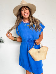 Woman in a blue dress and straw hat holding a beige bag against a white wall.
