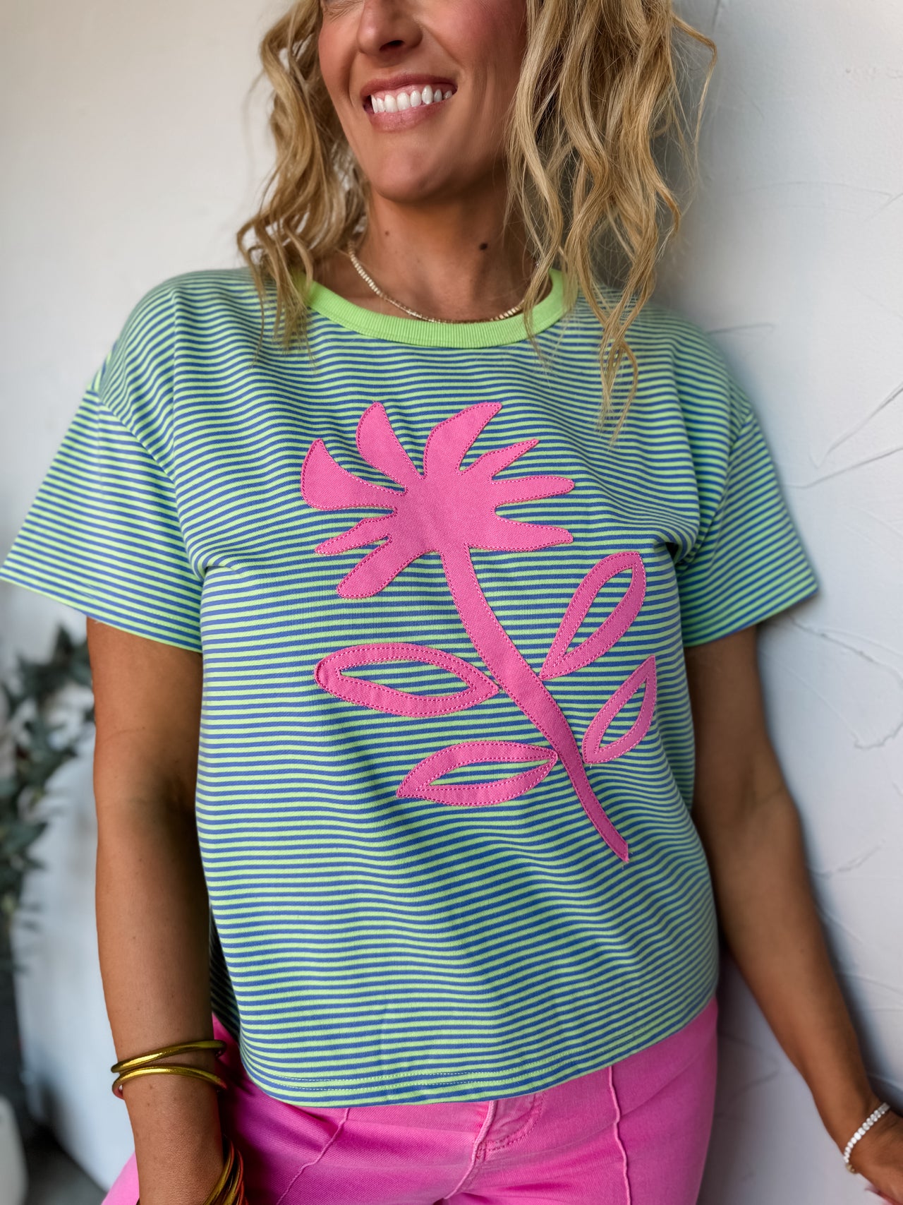 A smiling woman with wavy blonde hair leaning against a textured off-white wall, wearing a short-sleeve striped graphic t-shirt in the file 601CF9AF-AAB2-460F-BBFE-6E53DF56869E.jpeg. The relaxed-fit tee is crafted from a knit fabrication blended from 40% cotton, 54% polyester, and 6% rayon, featuring an all-over fine horizontal pinstripe pattern in bright lime green and turquoise blue. It is styled with a solid lime green contrast ribbed crew collar and highlights a large, stitched-on pink flower and stem a