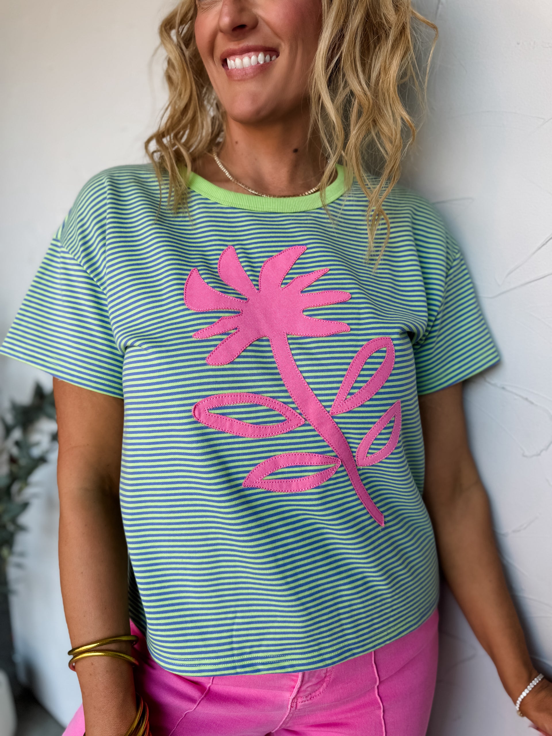 A smiling woman with wavy blonde hair leaning against a textured off-white wall, wearing a short-sleeve striped graphic t-shirt in the file 601CF9AF-AAB2-460F-BBFE-6E53DF56869E.jpeg. The relaxed-fit tee is crafted from a knit fabrication blended from 40% cotton, 54% polyester, and 6% rayon, featuring an all-over fine horizontal pinstripe pattern in bright lime green and turquoise blue. It is styled with a solid lime green contrast ribbed crew collar and highlights a large, stitched-on pink flower and stem a